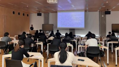 机に座る生徒たちとプロジェクタースクリーンがある教室の風景。